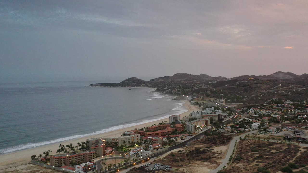 Aerial Los Cabos shoreline view with calm ocean water, white surf bands along the beach, clustered coastal buildings, a road corridor, low hills, and no visible people.