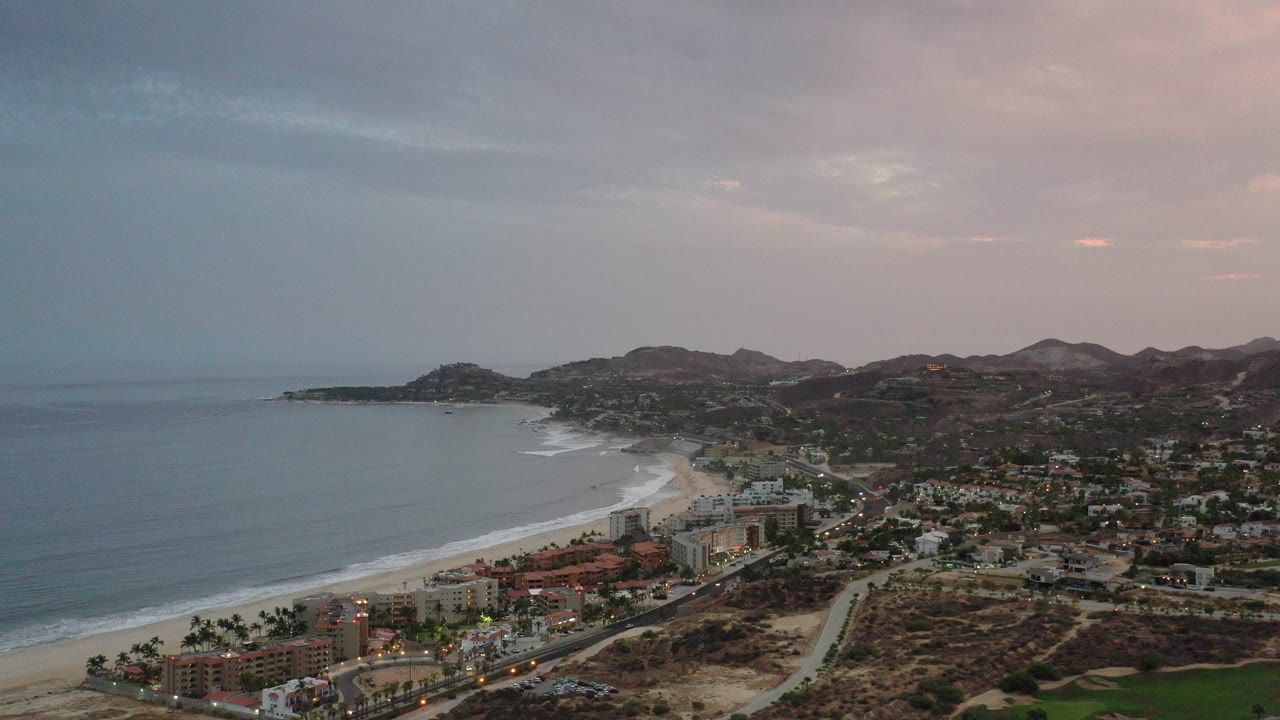 Wide aerial view over the Los Cabos coastline with the ocean on the left, a curving beach below, low hills in the distance, soft pink sky, and no visible people.