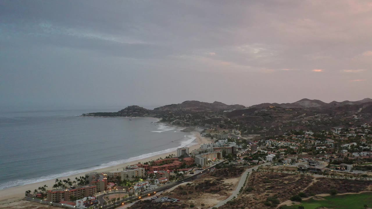 Aerial frame of Los Cabos with a broad ocean horizon, white surf along the beach, coastal buildings near the sand, dry hills beyond town, and no visible people.