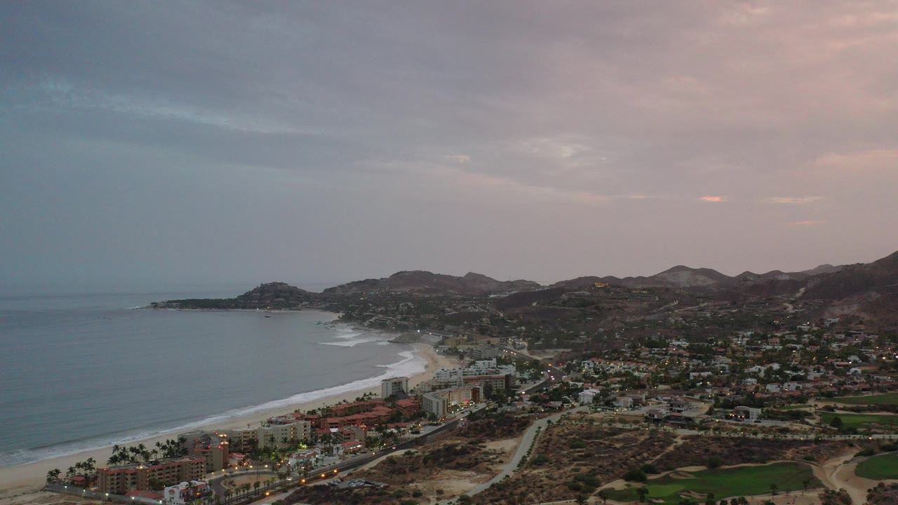 Wide aerial view of coastline, beach, town, and hills at dusk.