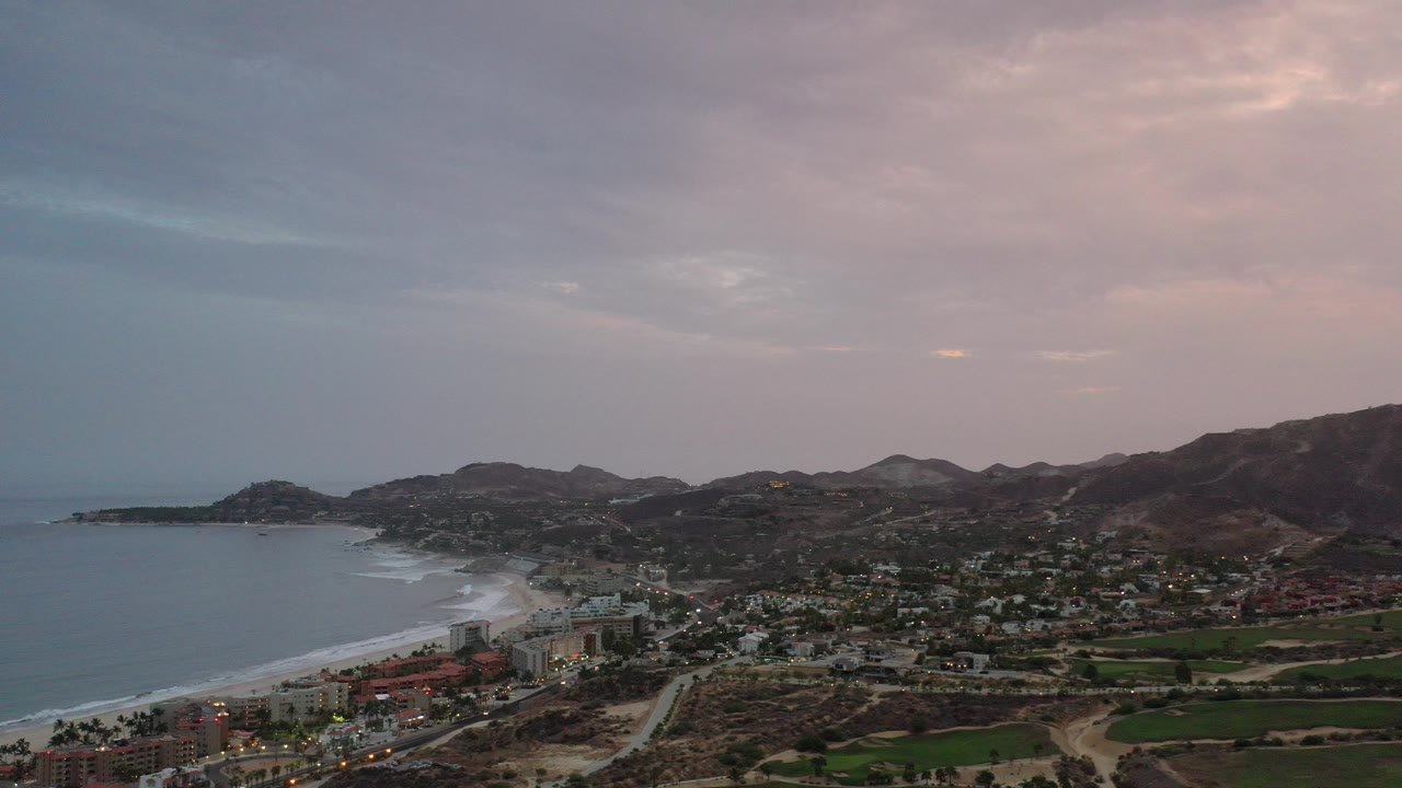 Los Cabos, Mexico, seen from an elevated viewpoint with ocean and beach at left, low buildings and roads below, dark hills to the right, and no visible people.