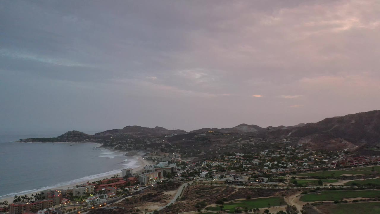 A wide Los Cabos coastal panorama shows the curving beach and surf on the left, town blocks and roads in the midground, mountain ridges beyond, and no visible people.