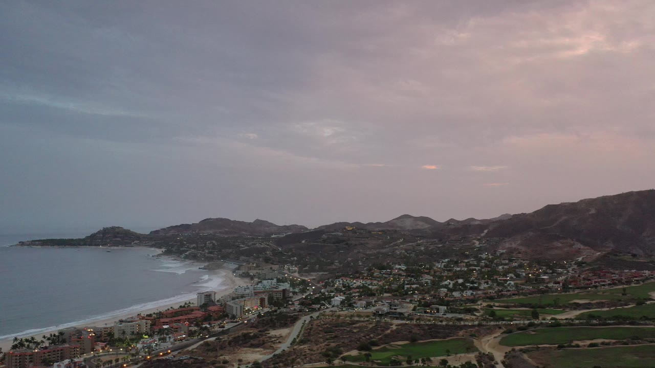 Beachfront buildings, roads, green fields, and mountains under evening clouds.