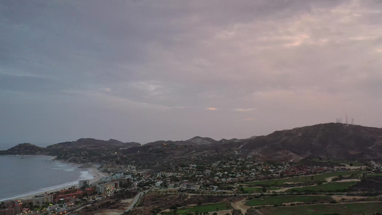 Early frame from the Los Cabos aerial view with bay water along the left edge, dense buildings near shore, green fairway-like patches, hill ridges, cloud cover, and no visible people.