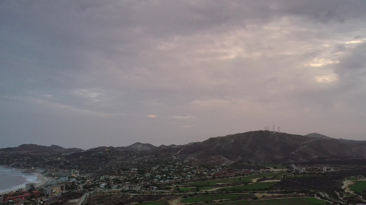 Opening frame of a Los Cabos, Mexico aerial view with low town buildings, green fields, distant water at the edges, rugged mountains, gray clouds, and no visible people.