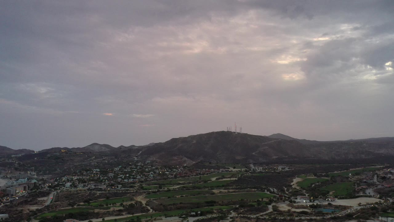 Wide aerial view of hills, town, roads, and green fields under cloudy sky.