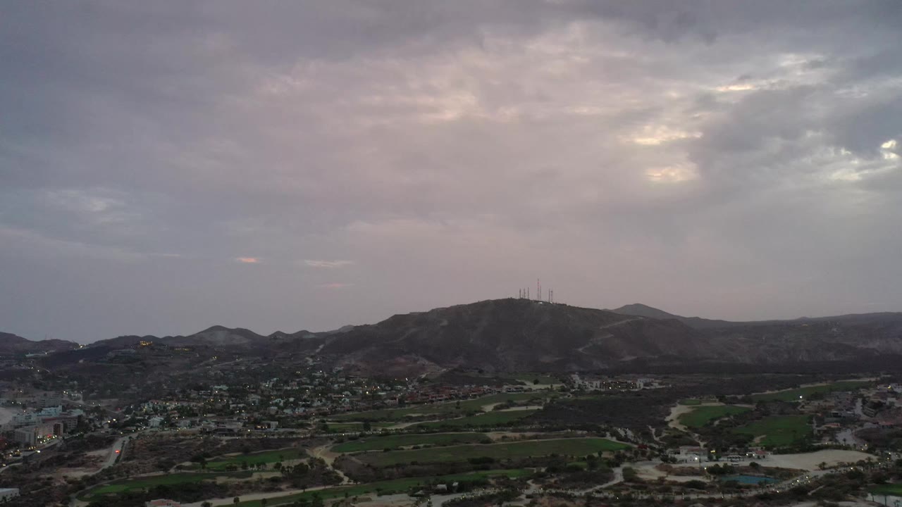 Los Cabos aerial view looking across roads, clustered buildings, green fields, and a hill with antenna structures below muted gray and pink clouds, no visible people.