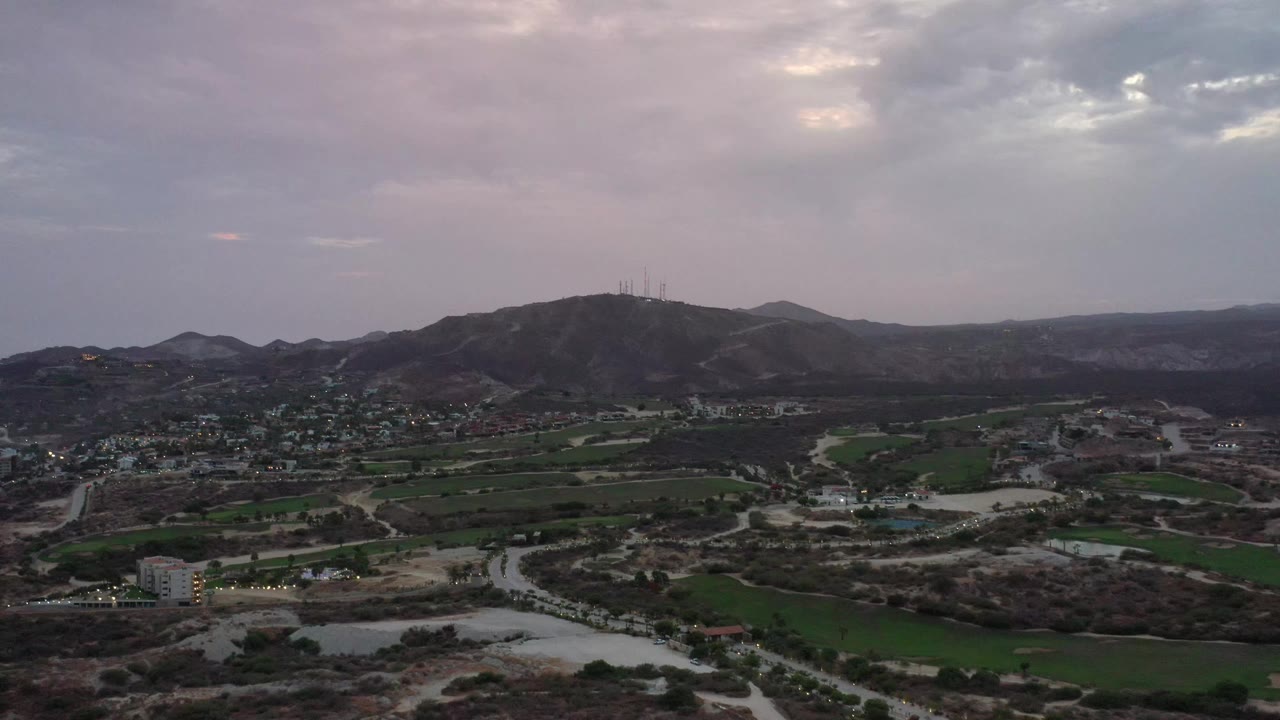Wide Los Cabos aerial frame with a hill and antenna structures centered beyond green plots, winding roads, scattered buildings, and no visible people.