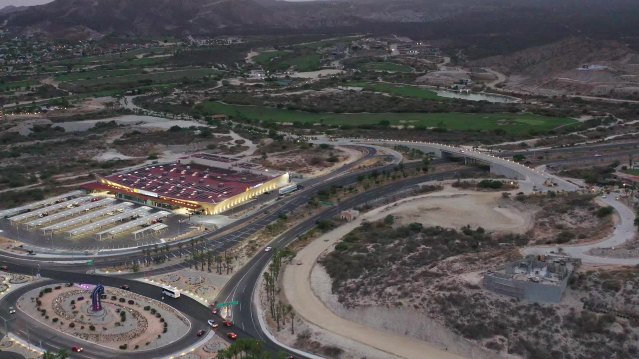 Aerial frame over Los Cabos roads with cars and a bus near the roundabout, a bright commercial structure to the left, open sandy lots, green fields, and mountains beyond.