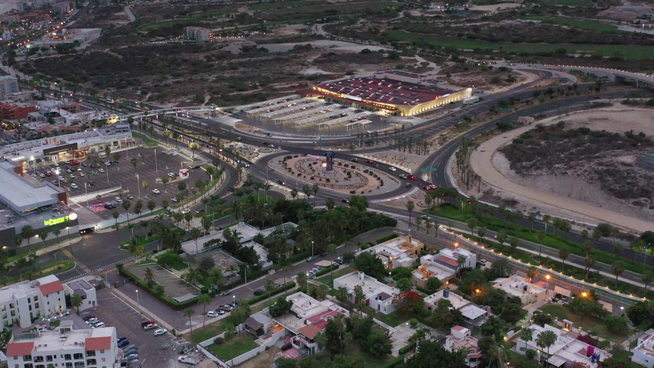 Oblique aerial view over Los Cabos, Mexico, showing curving roads, parking lots, low buildings, homes, and a lit commercial roof, with no visible people.