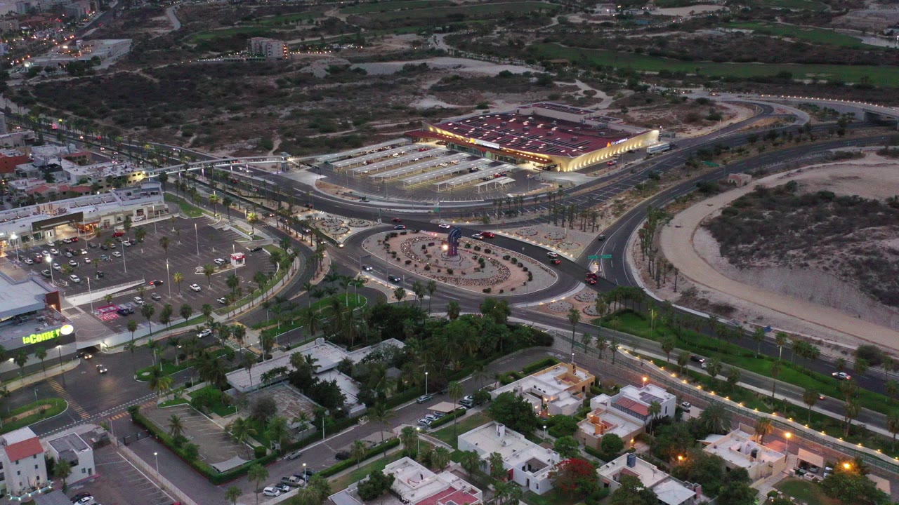 Aerial dusk view of a roundabout, roads, parking lots, and lit retail buildings.