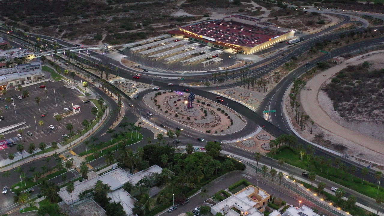 Aerial view in Los Cabos, Mexico, with curving roads, moving cars, parking areas, desert ground, and a lit commercial building near the top of frame, with no visible people.