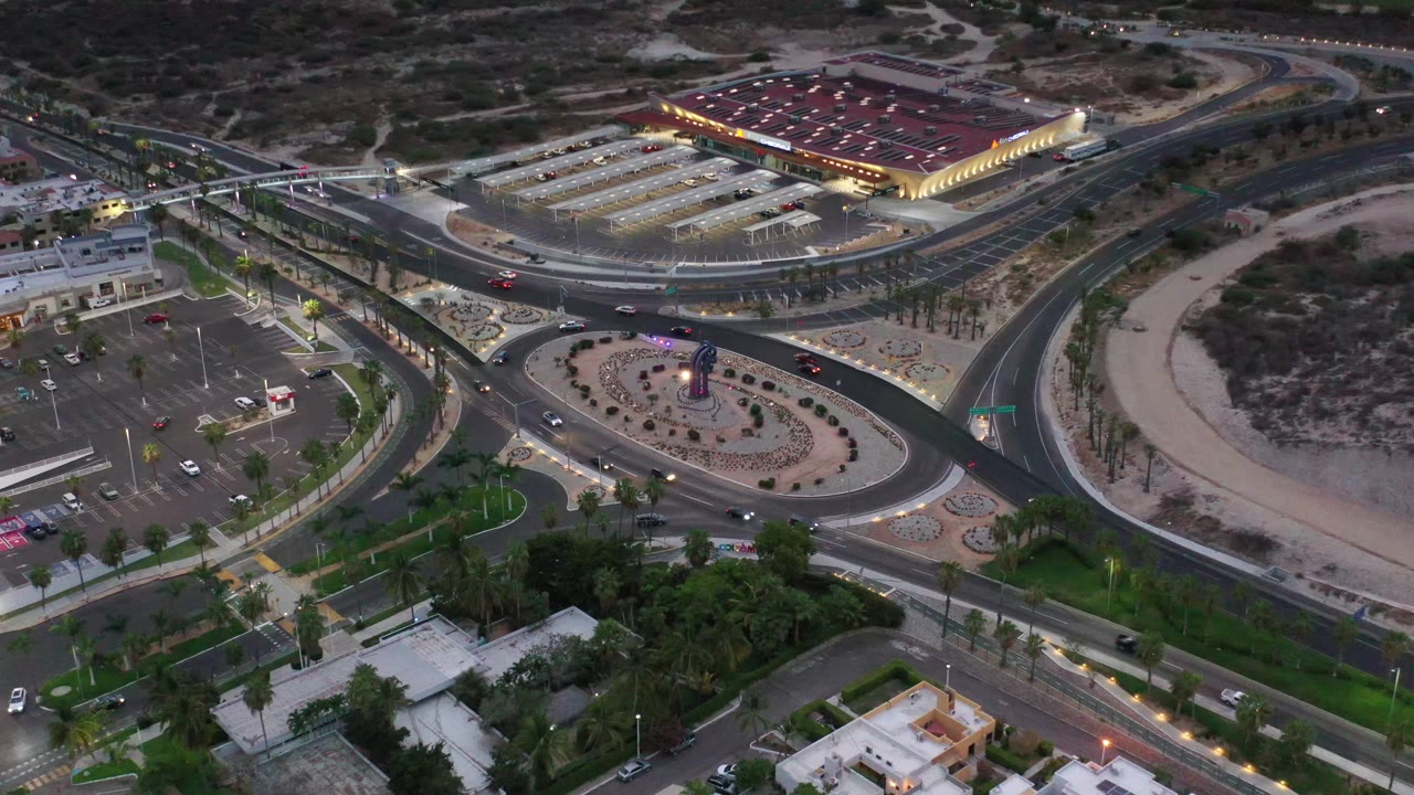Drone-height view over Los Cabos roads with cars spaced along the lanes, palm-lined landscaping, parking lots, and a brightly lit commercial building, with no visible people.