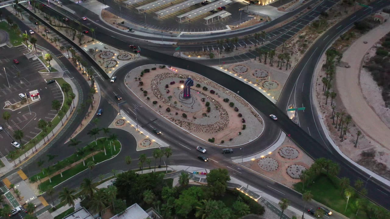 Aerial Los Cabos frame showing vehicles spaced across the curved lanes beside a long planted median, palm-lined walks, small parking areas, and no visible people.