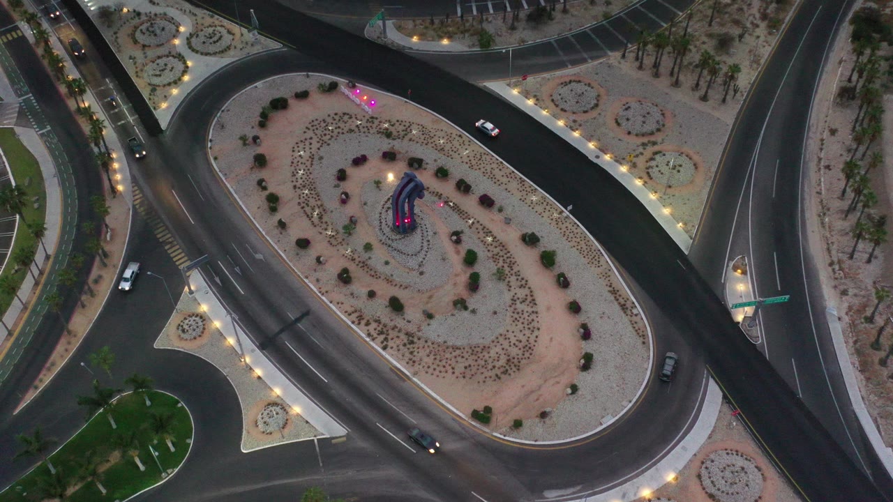 High overhead dusk view in Los Cabos, Mexico, showing a curved road around an oval desert median with shrubs, palms, streetlights, a lit sculpture, several cars, and no visible people.