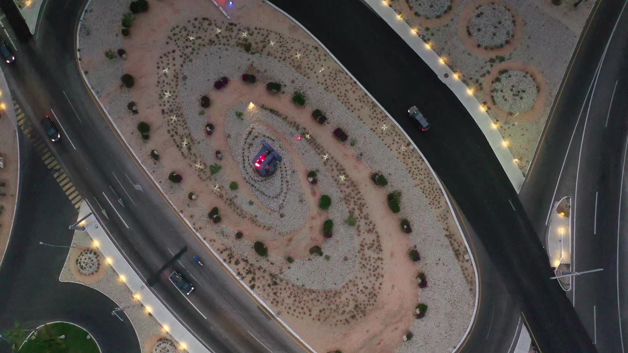 Overhead Los Cabos road scene with an oval landscaped island, dark traffic lanes, bright edge lights, a few cars on separate roads, and no visible people.