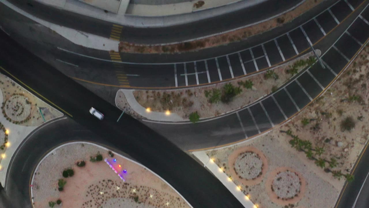 Aerial Los Cabos frame with a white car on the road, empty striped parking bays, planted desert beds, and no visible people.