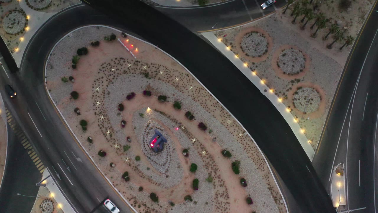 Overhead Los Cabos road scene with cars on multiple lanes, circular planting beds, illuminated sidewalks, and a pink-lit central feature, with no visible people.
