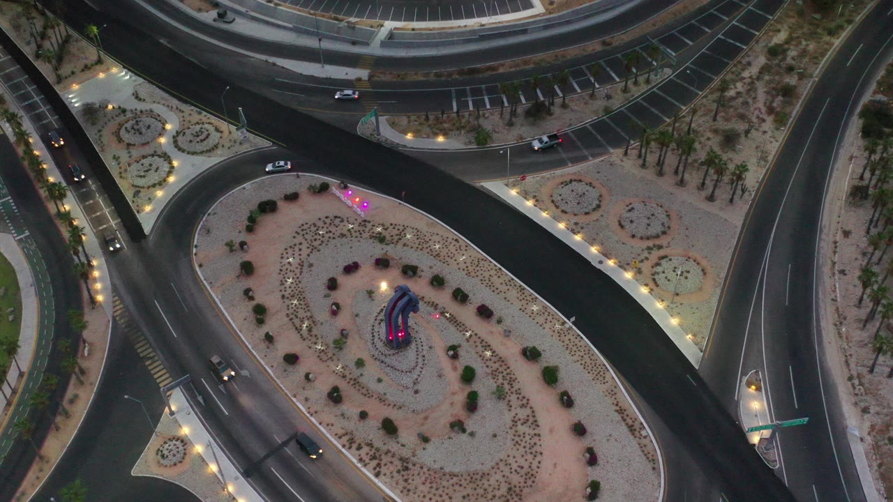 High overhead view in Los Cabos, Mexico showing a landscaped oval roundabout between curving roads, sparse cars, palms, path lights, and no visible people.