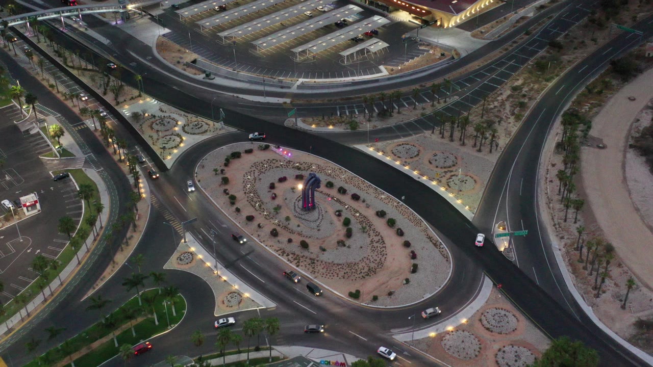 Top-down aerial frame in Los Cabos, Mexico, showing curved road lanes around a landscaped oval island, nearby parking areas, lit paths, and no visible people.