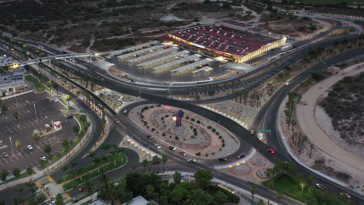 Opening aerial frame over Los Cabos, Mexico, with a landscaped roundabout below a large lit red-roofed building, surrounding parking lots, curving roads, desert scrub, and no visible people.