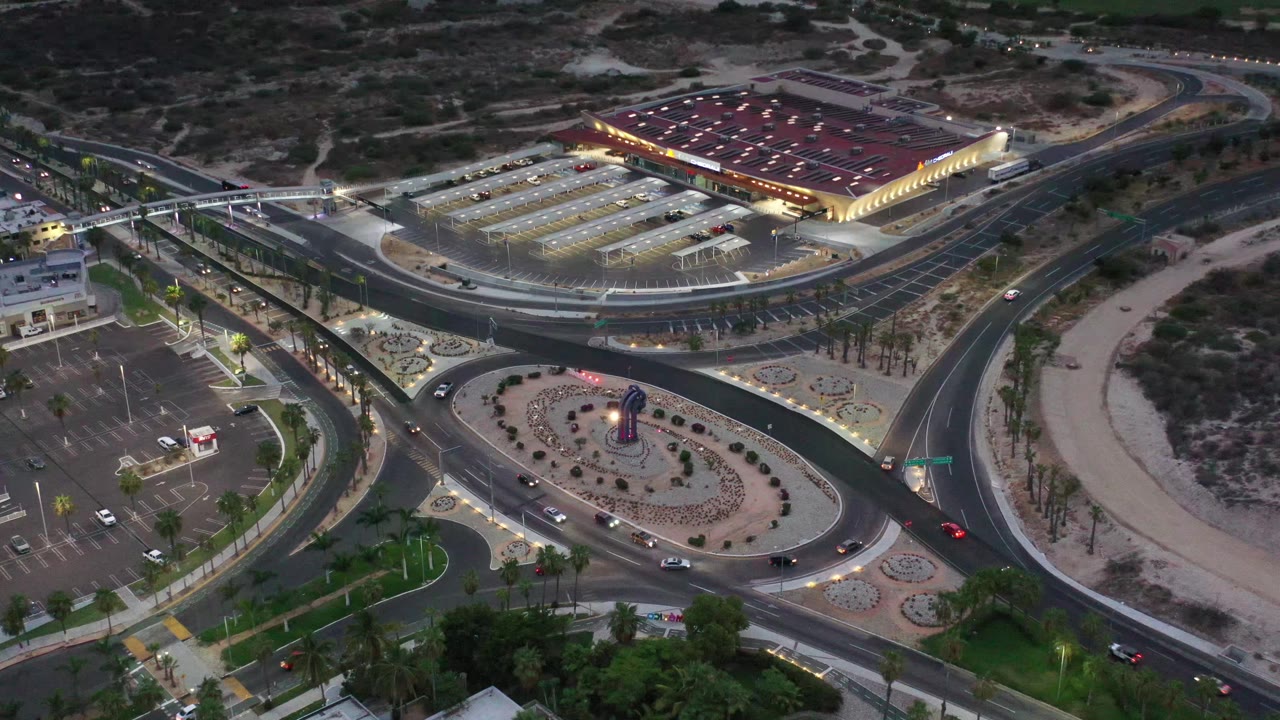 Aerial view in Los Cabos, Mexico, showing the roundabout slightly more centered, with cars on the curved roads, lit parking rows, dark desert terrain, palm landscaping, and no visible people.