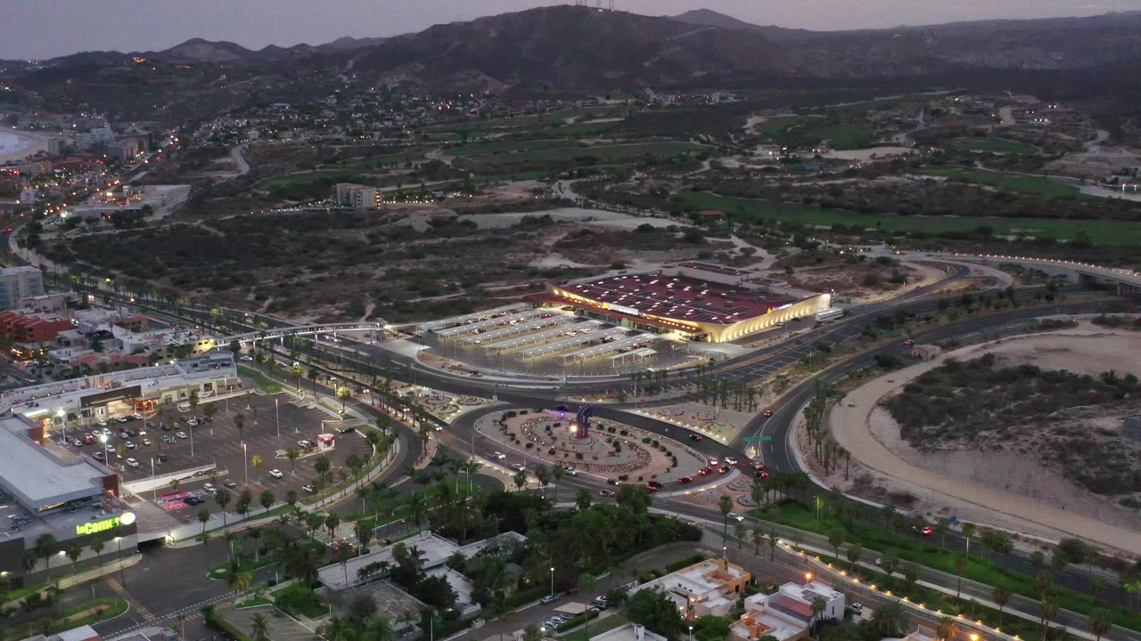 Aerial Los Cabos dusk frame centered on a bright retail complex, surrounding roads, parked cars, green field patches, dry lots, mountain ridges, and no visible people.