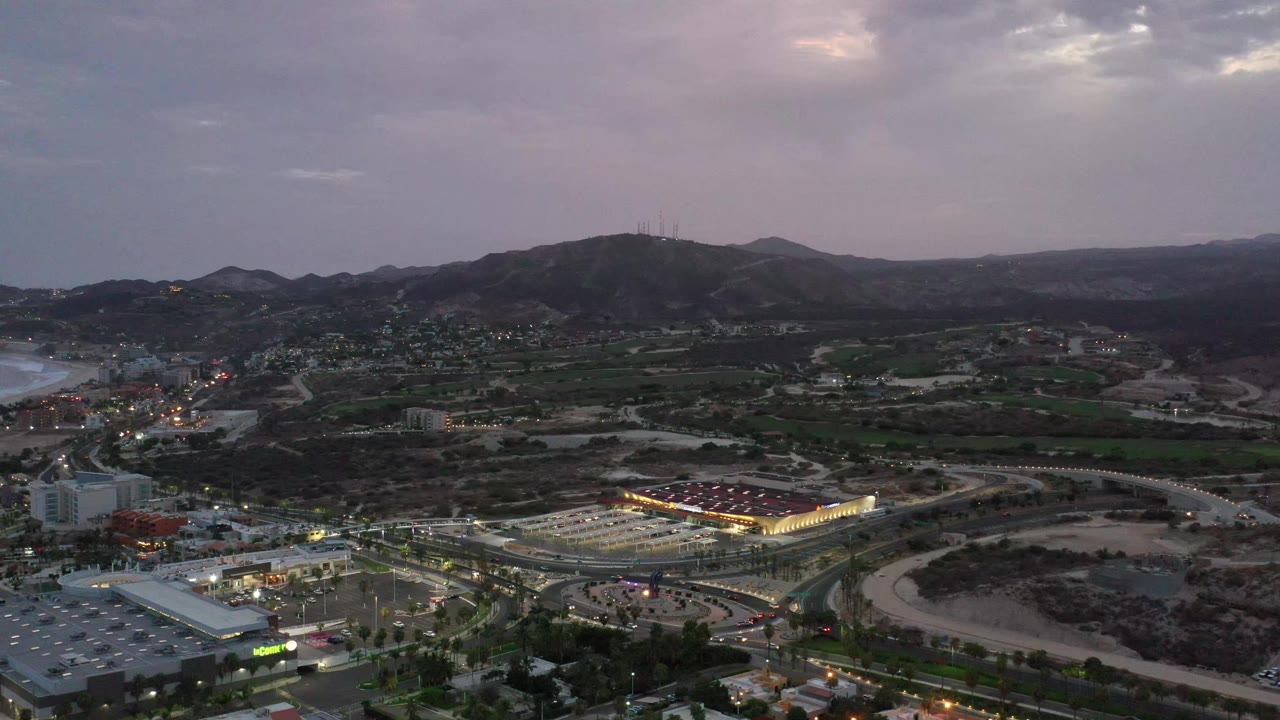 Los Cabos aerial view one second in, showing a bright rectangular building cluster, curving roads, dark hills, distant settlement lights, and no visible people.