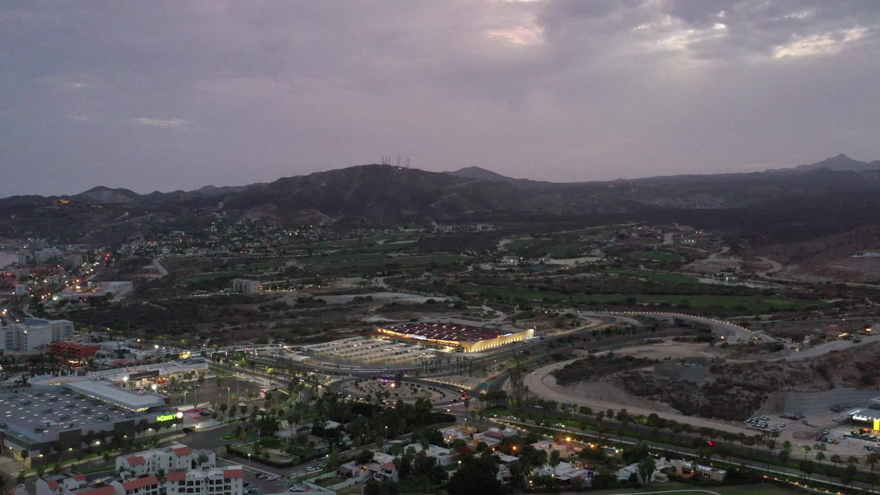 Wide aerial frame over a Los Cabos resort area at evening, with lit buildings and roads below dark hills under a purple gray sky, no visible people.