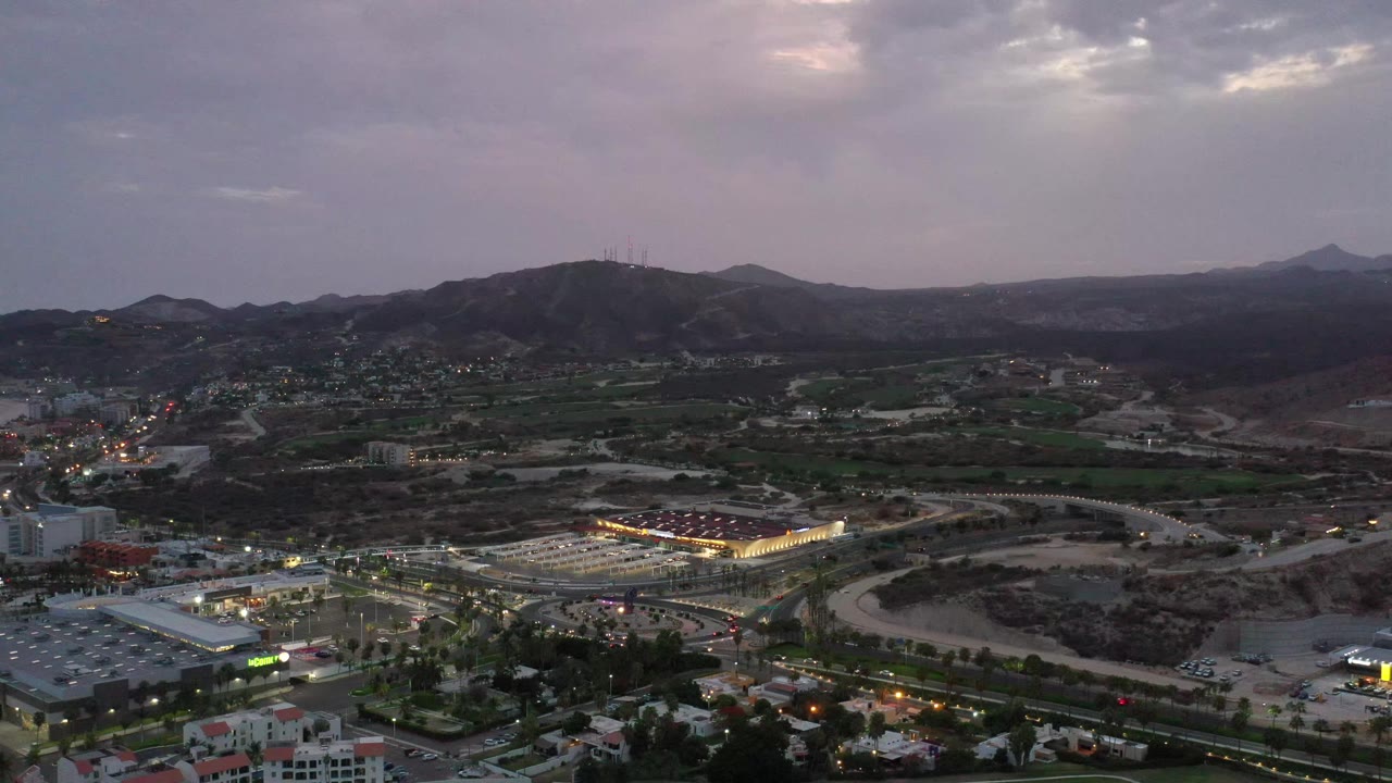 Los Cabos evening aerial with a bright rectangular resort complex near the lower center, curved roads, scattered lights, and mountain ridges in the distance, no visible people.