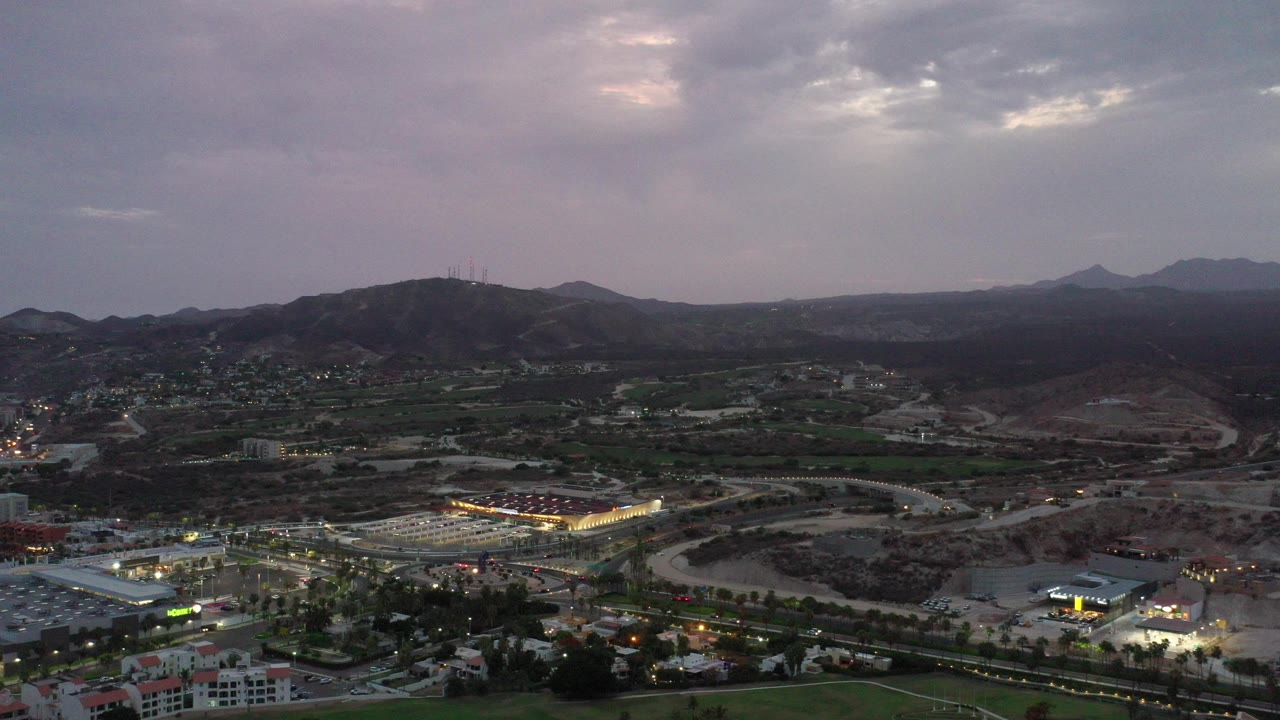Wide aerial view over Los Cabos, Mexico, with roads, low buildings, green grounds, rocky hills, distant mountains, purple gray clouds, and no visible people.