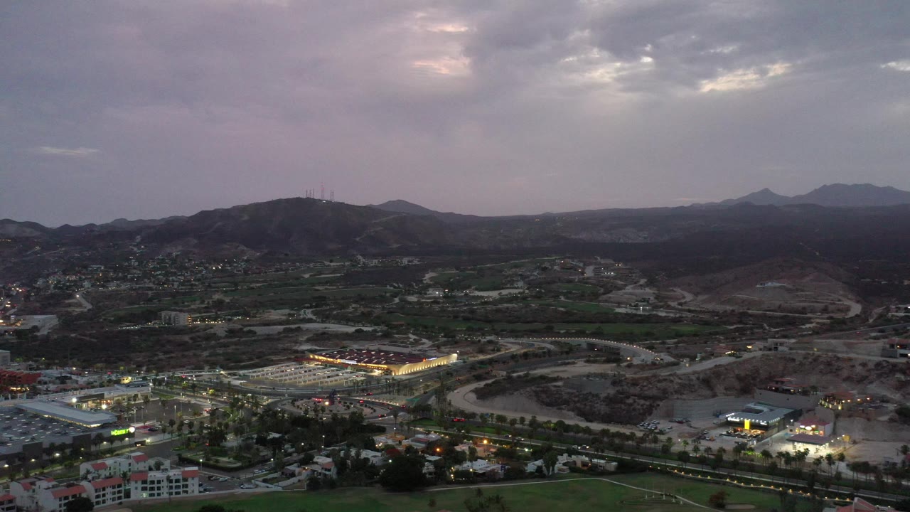 Wide dusk aerial over roads, buildings, hills, and layered mountains.