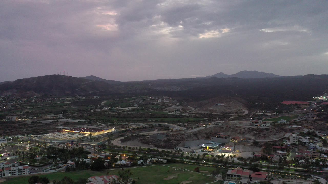 Wide aerial view over Los Cabos, Mexico with low buildings, roads, dark mountain ridges, and a cloudy gray-purple sky, no visible people.