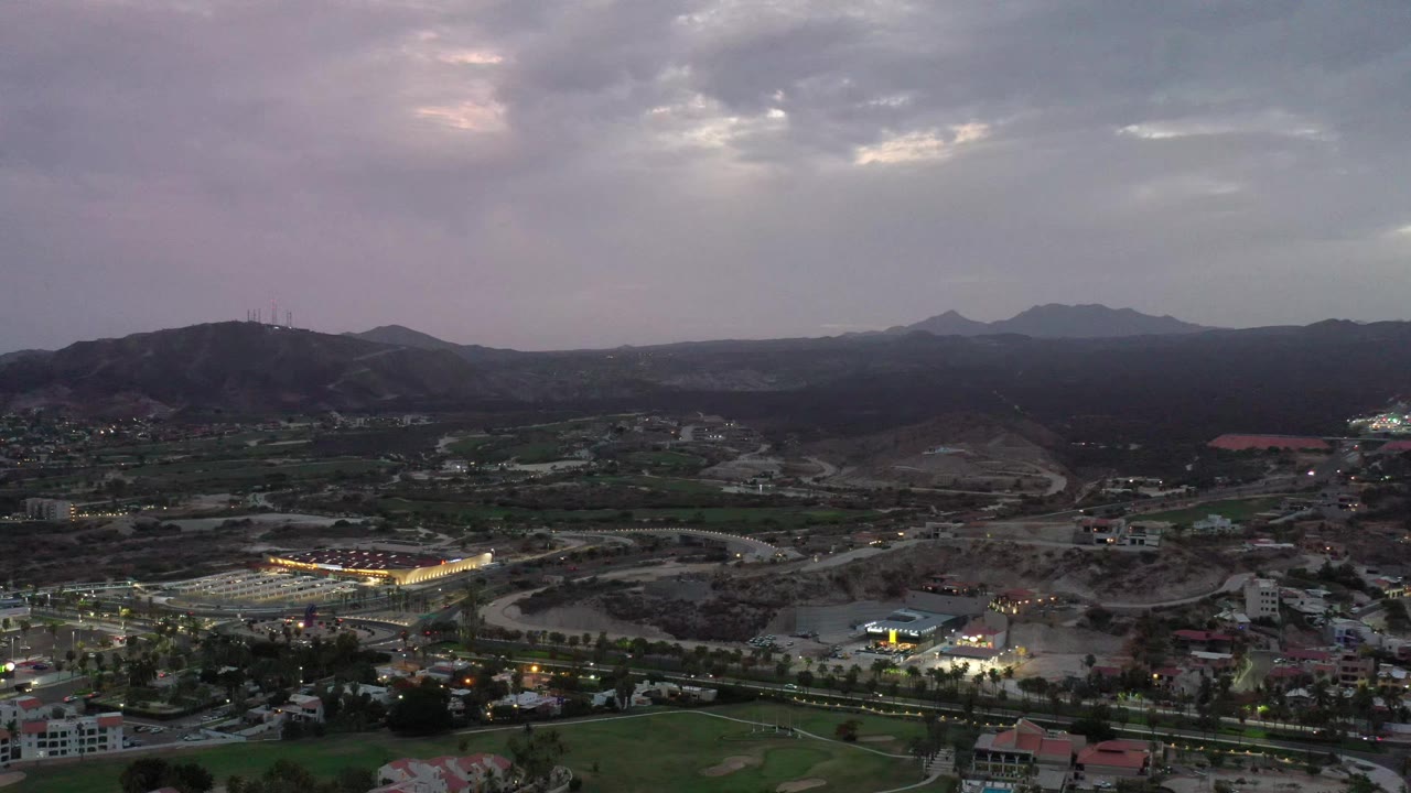 Elevated Los Cabos view with bright building lights in the lower left, winding roads through the valley, mountain silhouettes, and heavy clouds, no visible people.
