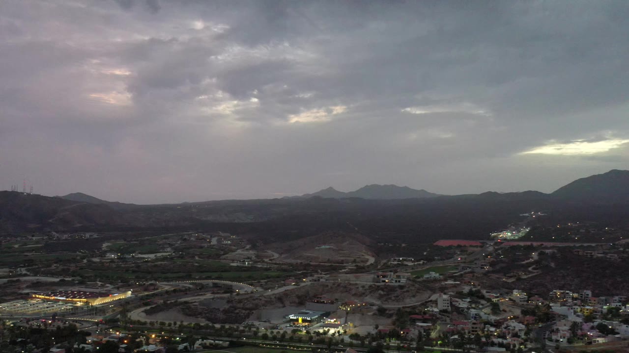 Los Cabos, Mexico aerial frame with the horizon stretching across dark hills, a pale break in the clouds, roads below, and no visible people.
