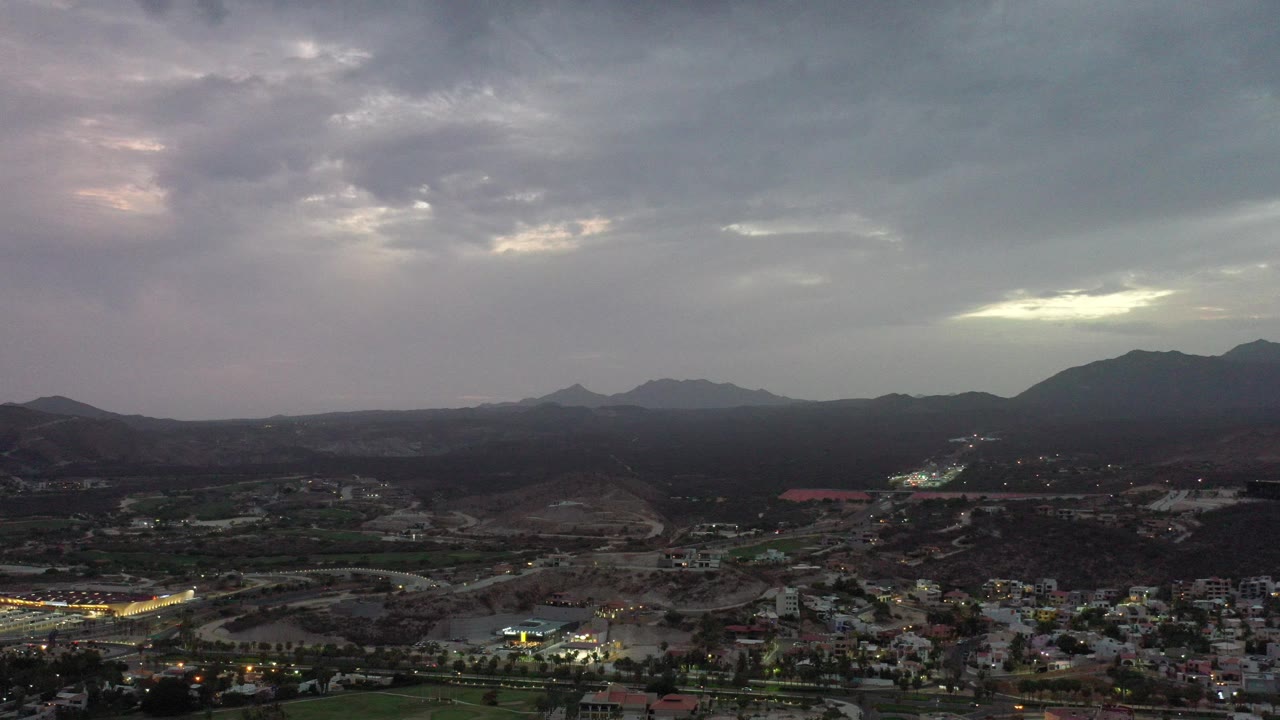 Wide aerial view in Los Cabos, Mexico, with gray clouds above dark mountain ridges, town buildings and roads below, and no visible people.