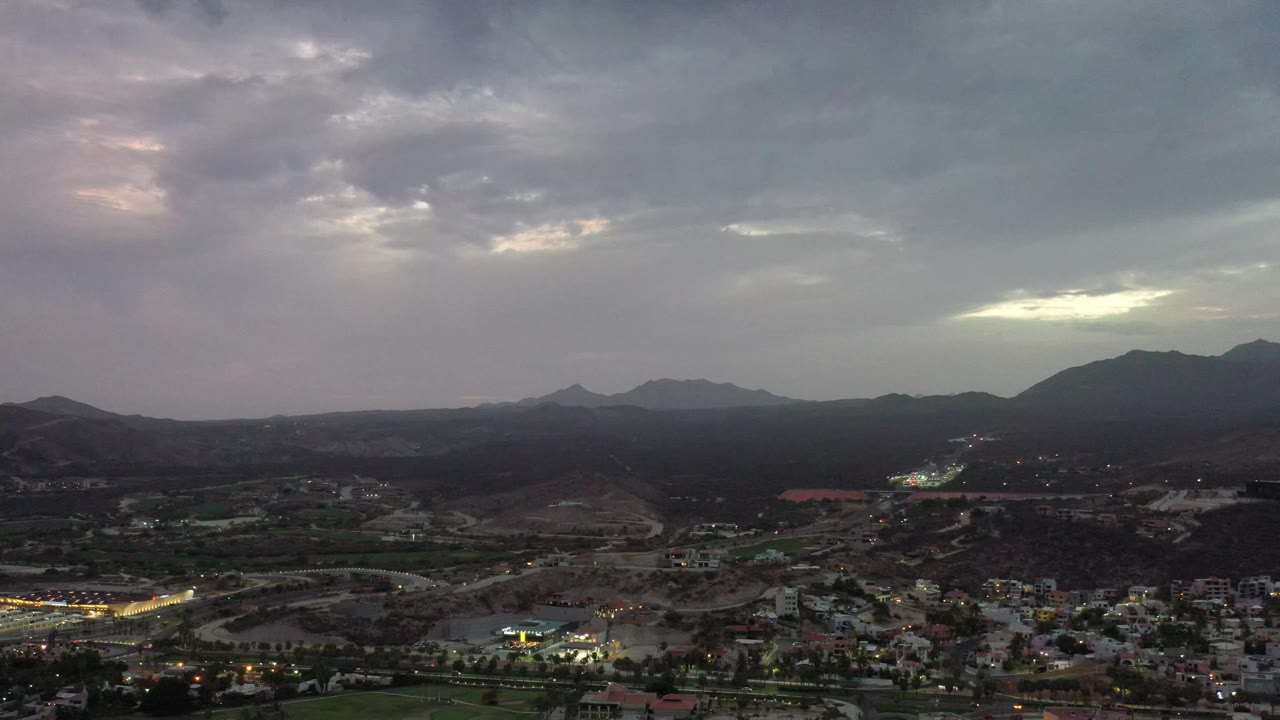 Los Cabos aerial frame with a dark hillside midground, a small cluster of buildings and lights at the bottom, and a pale opening in the cloud deck, with no visible people.