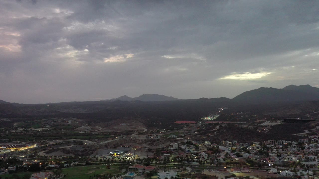 Wide aerial view of a town below dark hills and cloudy sky.