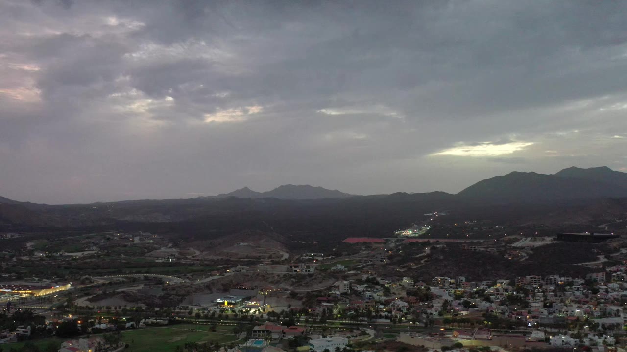Aerial view across Los Cabos, Mexico, showing clustered buildings at the lower edge, rolling dark terrain, mountain silhouettes, and no visible people.