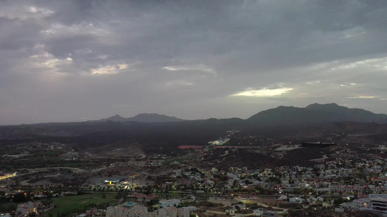 Los Cabos, Mexico is seen with low urban blocks, lit roads, dark surrounding hills, and a pale opening in the cloudy sky, with no visible people.