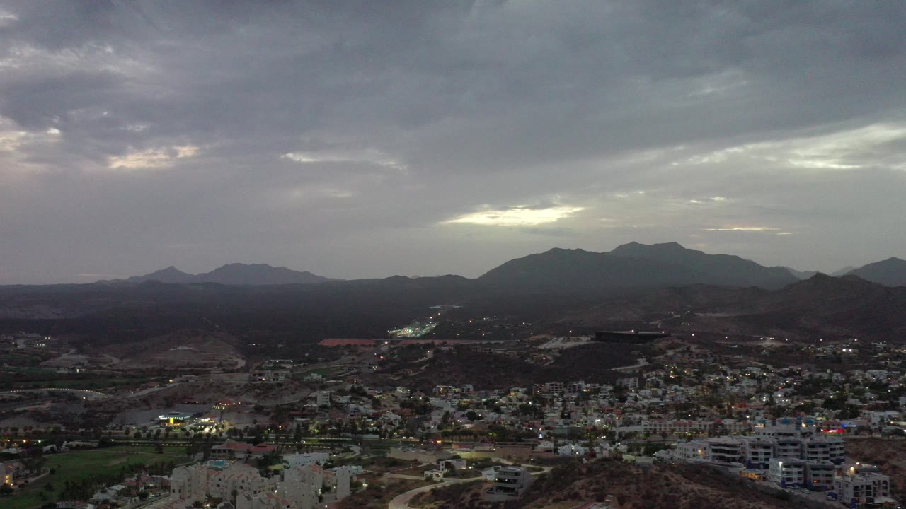 Wide aerial view over Los Cabos, Mexico, with a city in the lower frame, dark mountain ridges on the horizon, gray clouds overhead, and no visible people.