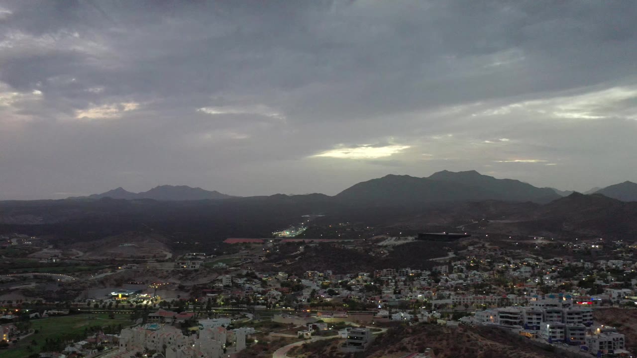 Aerial Los Cabos cityscape with low buildings and roads in the foreground, dark hills across the middle distance, a muted mountain skyline, and no visible people.