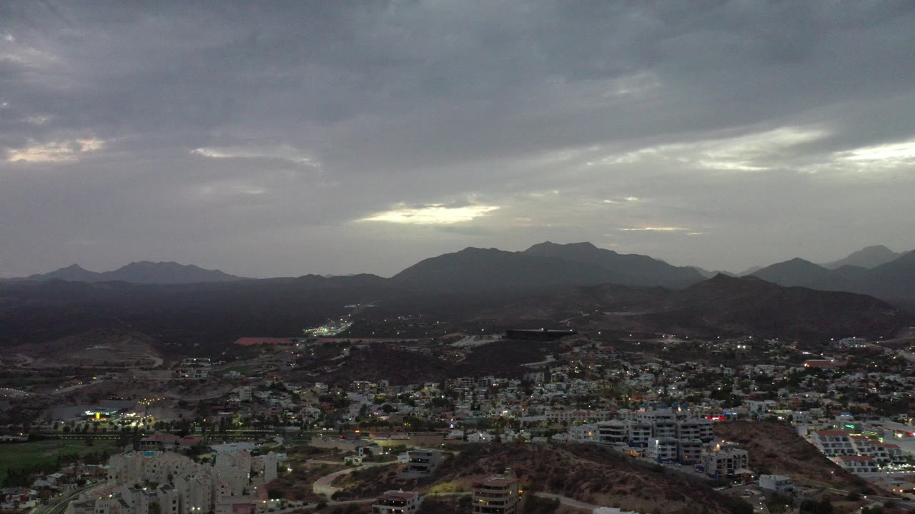 Los Cabos, Mexico, appears from an elevated distance with city buildings in the lower frame, dark hills beyond, gray cloud cover above, and no visible people.