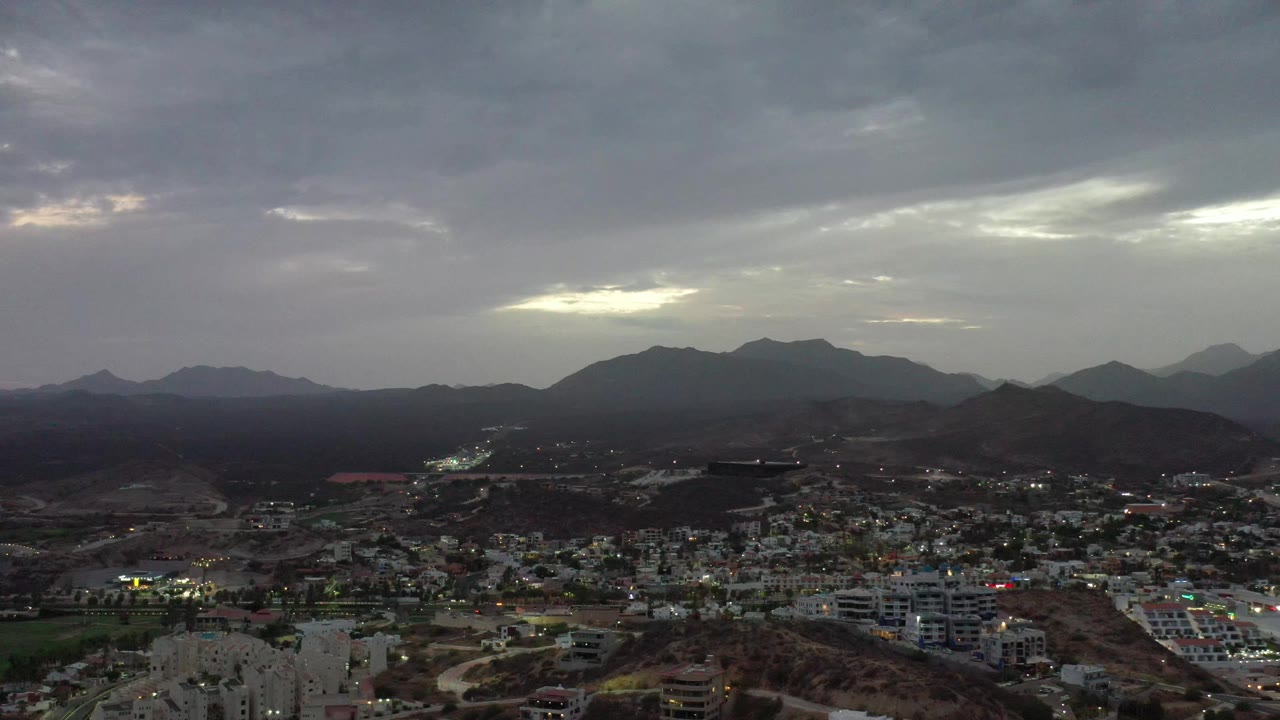 The Los Cabos aerial view holds on rooftops and streets below layered dark hills, with a brighter opening in the cloud cover and no visible people.