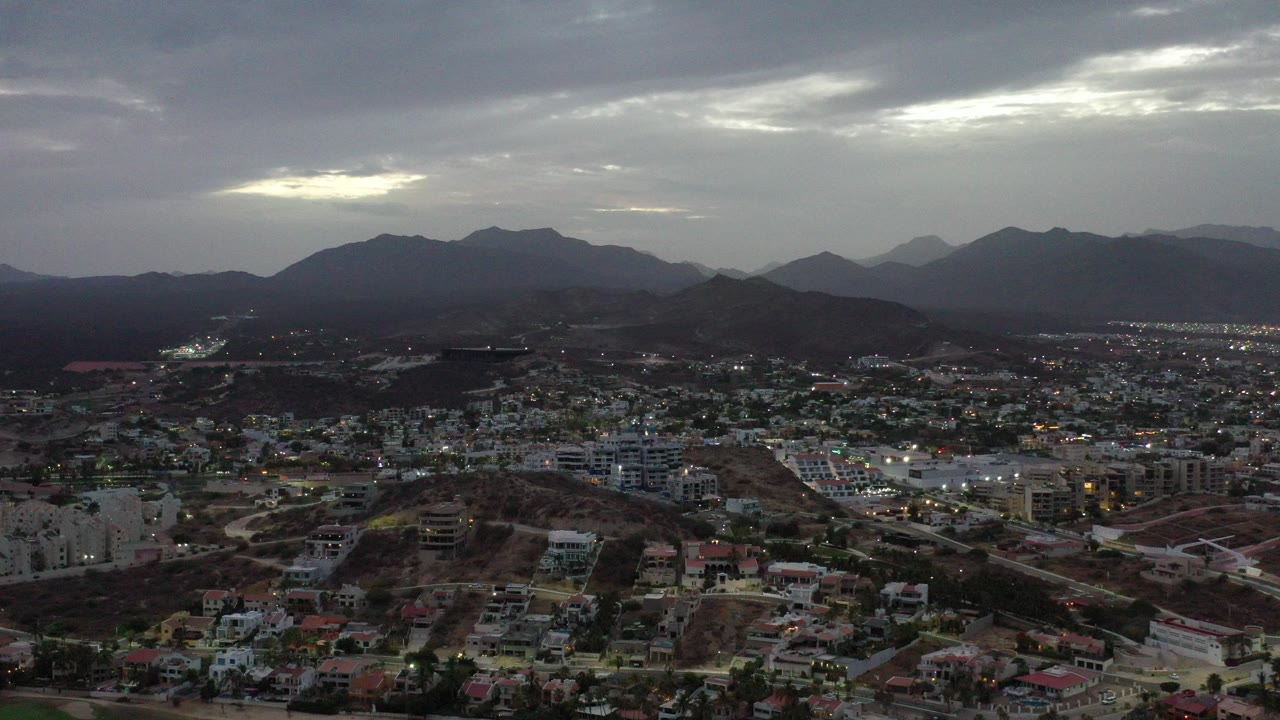 Opening frame of Los Cabos, Mexico seen from above, with low-rise buildings, roads, dark hills, distant mountains, gray cloud cover, and no visible people.