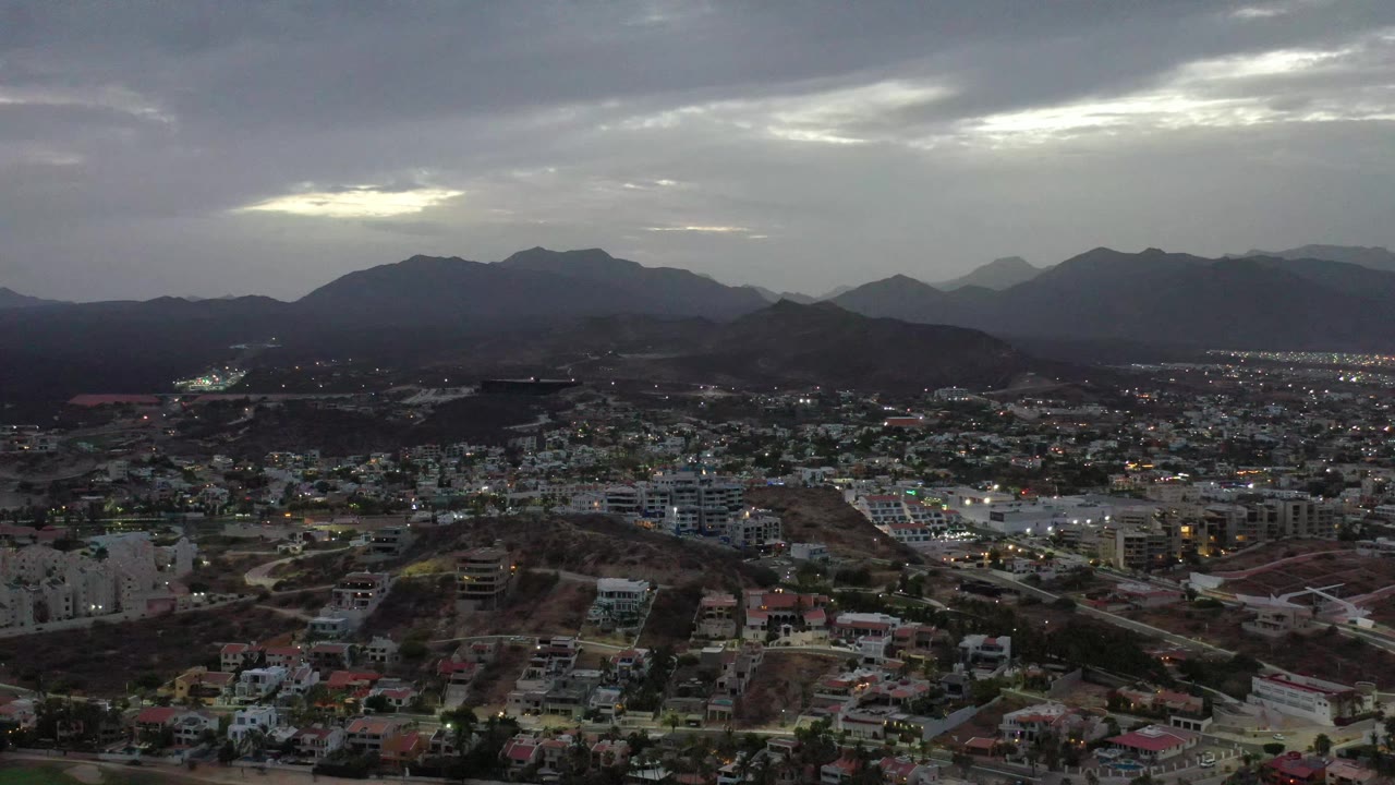 A second high view over Los Cabos, Mexico shows clustered buildings and streets below a dark mountain ridge, with bright breaks in the gray clouds and no visible people.