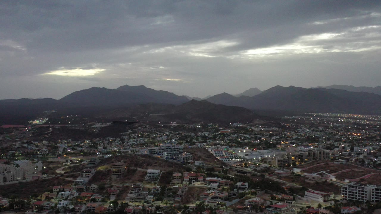 Wide elevated view of Los Cabos, Mexico with suburban buildings below, dark mountain silhouettes on the horizon, gray cloud cover above, and no visible people.