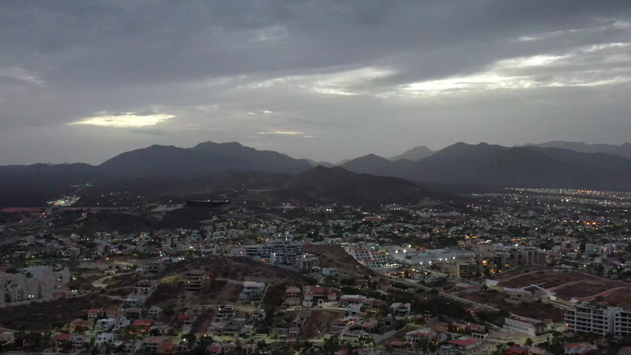 Los Cabos, Mexico appears from an elevated position with clustered buildings in the lower frame, layered hills beyond, a broad gray sky, and no visible people.