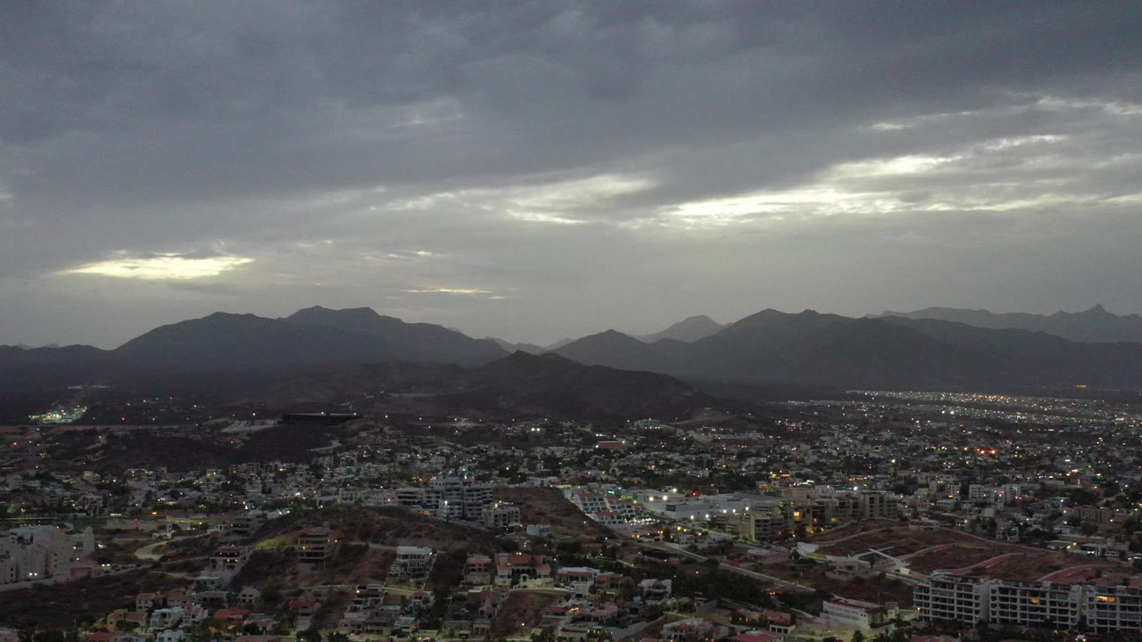 Wide aerial view of houses, hills, mountains, and heavy clouds at dusk.