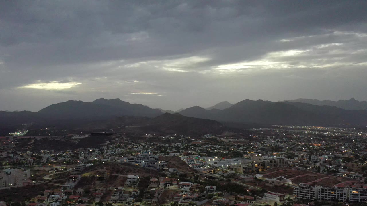 Los Cabos, Mexico appears from above with dense housing, scattered lights, layered hills, distant mountains, and a brighter opening in the cloud cover, with no visible people.